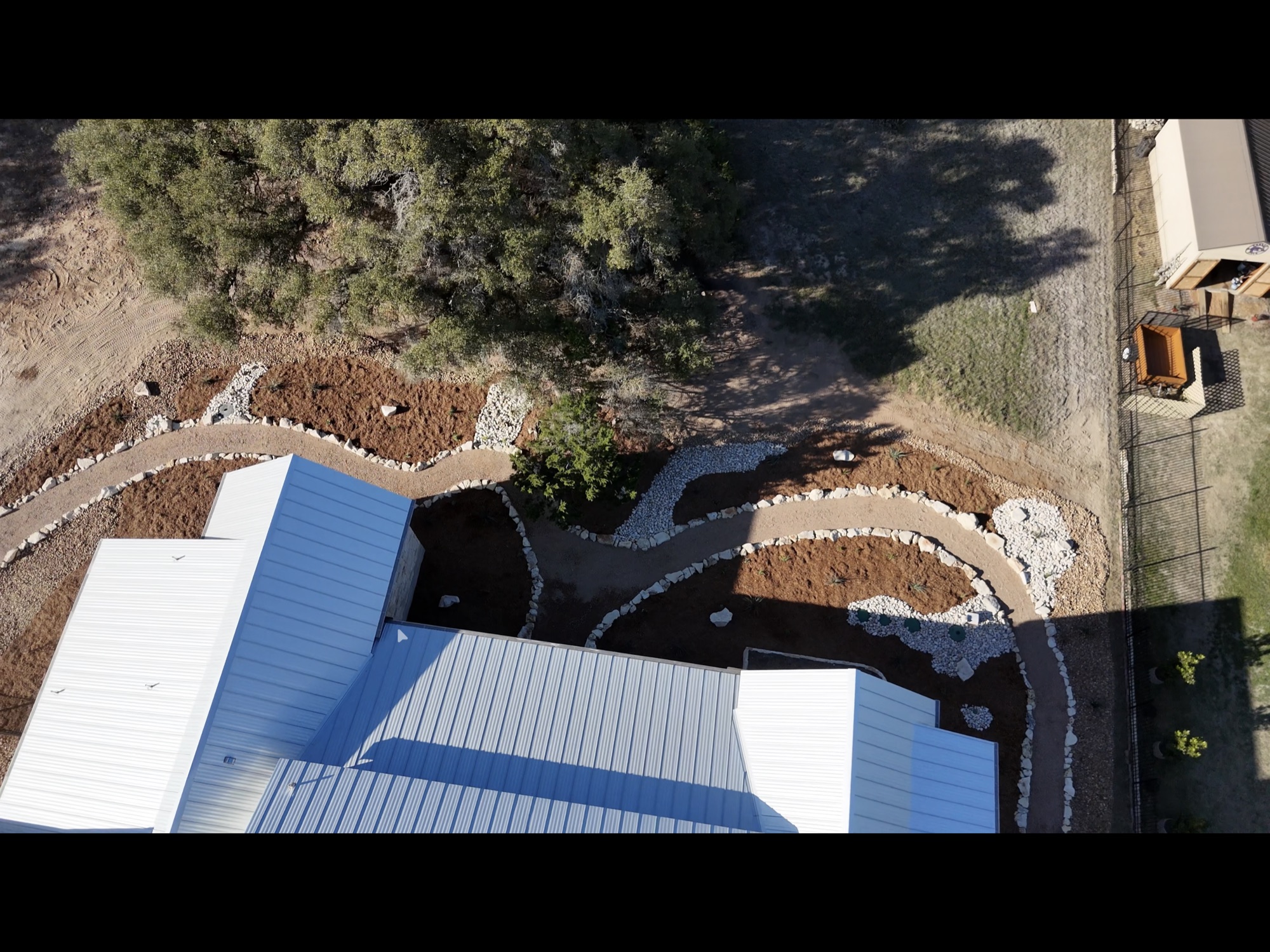 Aerial of finished native bed work — curving limestone-bordered beds, decomposed granite paths, mulch, around a metal-roof outbuilding